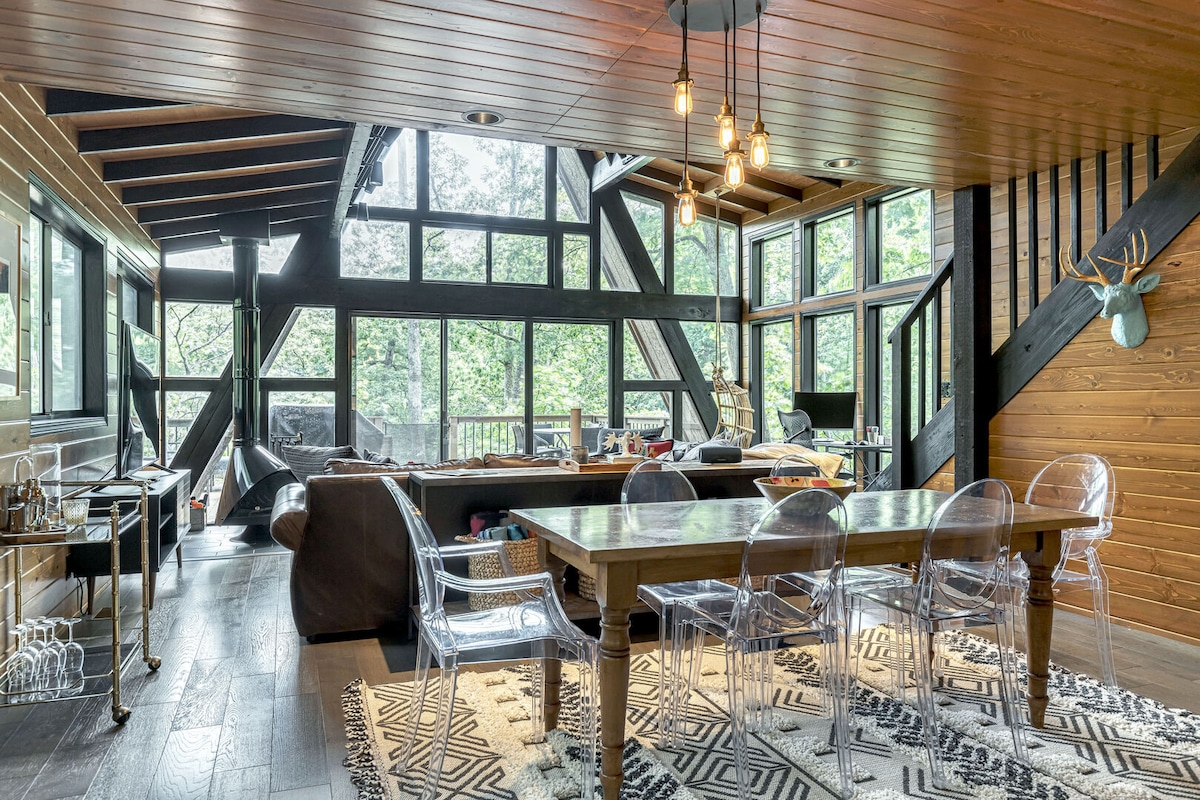 A spacious open living area showcases large A-frame windows, inviting natural light and views of the surrounding trees. A wooden dining table with transparent chairs is positioned in the foreground, while a comfortable seating area is glimpsed in the background, adorned with a cozy rug.