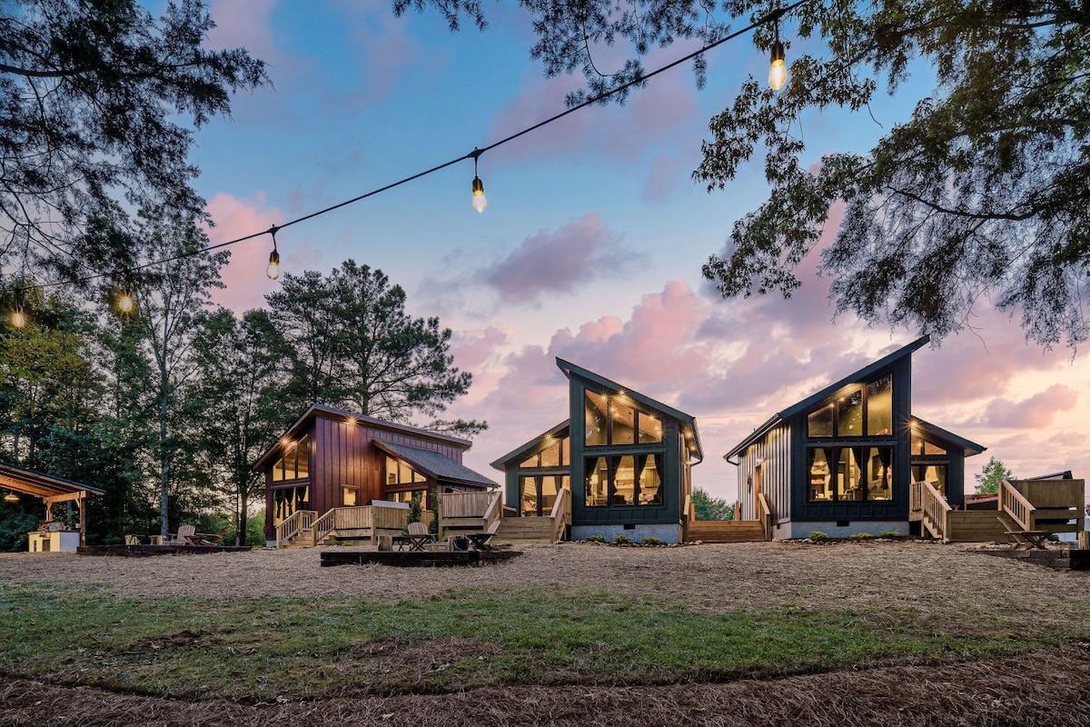 Three modern cabins are positioned together, featuring large glass windows and unique rooflines. An outdoor seating area with wooden chairs is visible in front, set against a backdrop of trees and a colorful sky at dusk, enhanced by string lights overhead.
