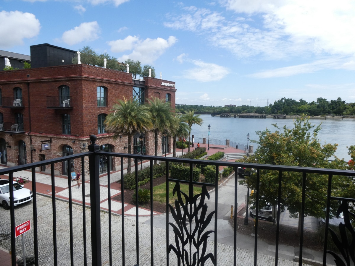 A panoramic view from a private balcony showcases the Savannah River, lined with lush palm trees and walking paths. The historic brick building is complemented by clear blue skies and soft clouds, while cobblestone streets provide texture in the foreground.