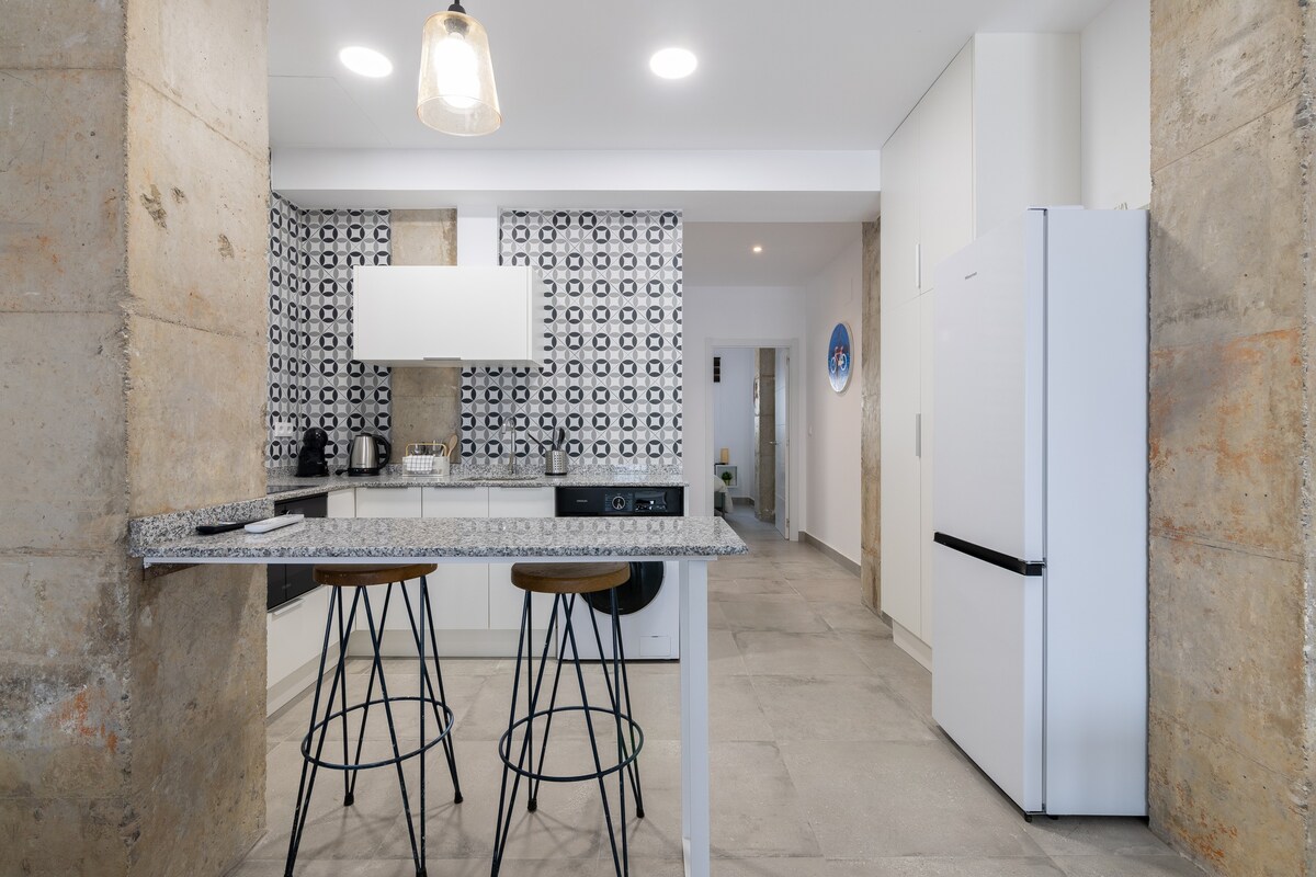 A modern kitchen space is showcased, featuring a bar area with two tall stools. The backsplash is adorned with a distinctive black and white pattern. A spacious countertop and white cabinetry complement the overall design, providing a clean and functional atmosphere.