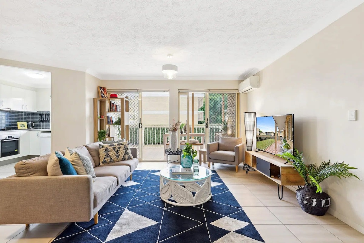 A bright living area features a comfortable sofa adorned with colorful cushions, paired with a round coffee table. A flat-screen TV is positioned on a wooden console, while large glass doors open to an outdoor space. A geometric area rug adds a modern touch to the tiled floor.