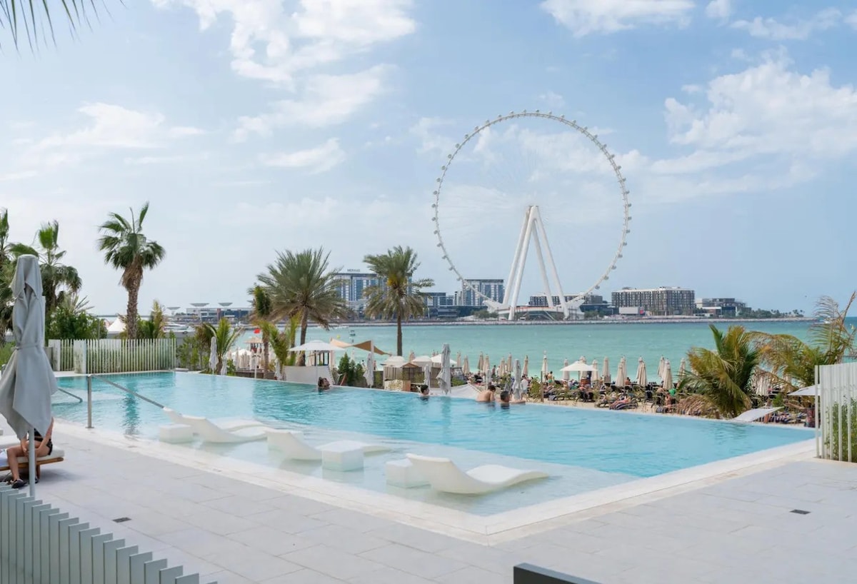 An infinity pool features comfortable loungers positioned beside it, overlooking a vibrant beach scene. In the background, the iconic Ferris wheel stands against a blue sky, framed by palm trees and beach umbrellas.