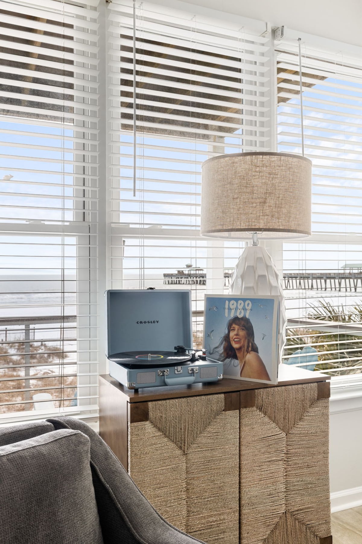 A coastal-inspired living area features a turntable atop a textured cabinet, accompanied by a music album placed nearby. Natural light filters through the blinds, illuminating the ocean view outside the large windows, which frame the scene with a soft ambiance.