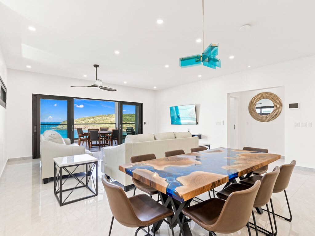 A modern dining area is highlighted by a striking wooden table with blue resin inlays. Comfortable chairs surround the table, and large glass doors open to reveal expansive views of the ocean and surrounding landscape, allowing natural light to fill the space.