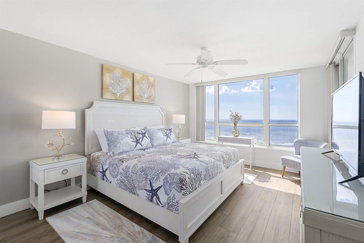 A spacious bedroom features a large bed with a blue and white ocean-themed quilt. Natural light floods the room through expansive windows showcasing views of the Gulf of Mexico. A cozy chair and small bedside table complement the elegant decor, creating a serene retreat.