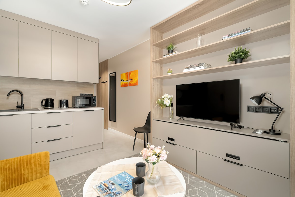 A modern studio apartment features a well-equipped kitchenette with sleek cabinetry, a microwave, and coffee-making facilities. A comfortable seating area includes a mustard-colored sofa and a coffee table with flowers. A SmartTV on a contemporary console offers entertainment options, and decorative shelves add charm.