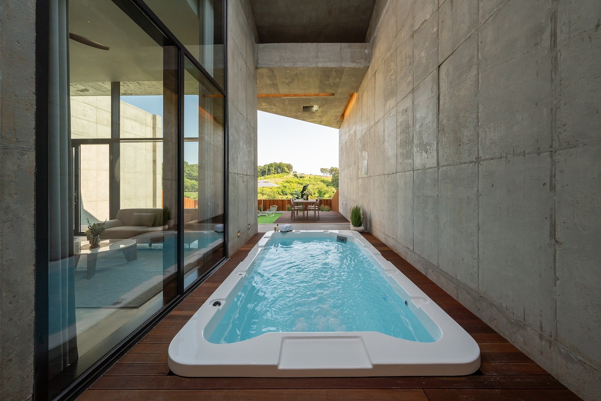 An indoor spa area is highlighted, featuring a white hot tub filled with clear water. Concrete walls and large glass doors create a seamless indoor-outdoor flow. Natural light illuminates the space, showcasing surrounding greenery visible through the open doors.