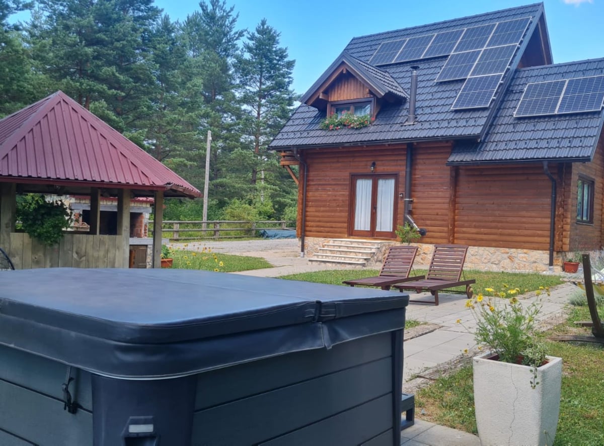 A charming wooden house is framed by lush greenery and features a sloped roof. Solar panels are installed on the roof. A hot tub sits in the foreground, with two lounge chairs positioned on a paved area. A gazebo can be seen adjacent to the house.