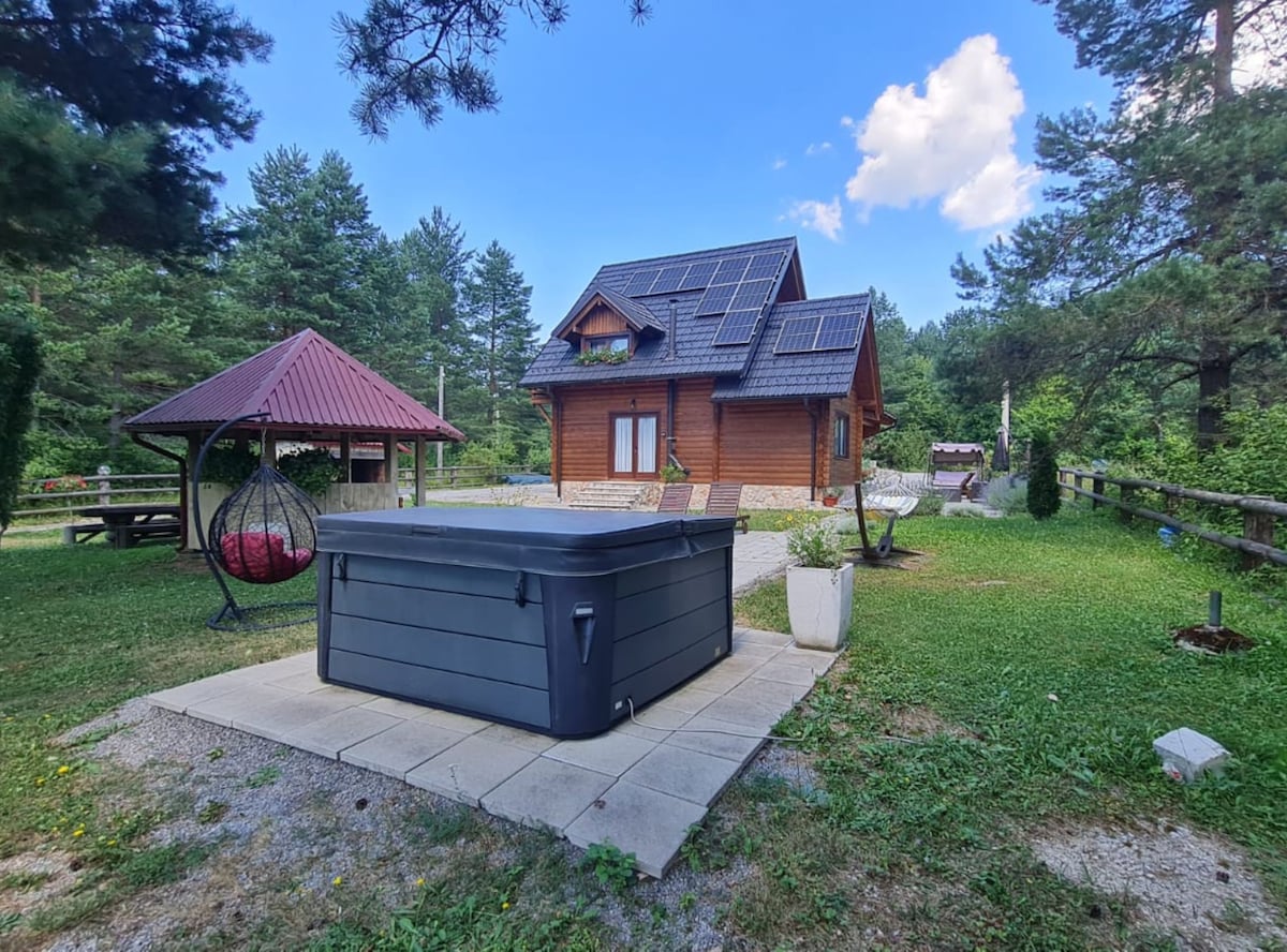 A wooden cabin is set among lush greenery, featuring solar panels on the roof. A hot tub is positioned on a tiled patio in the foreground. A shaded gazebo with a red roof and nearby picnic area are visible, surrounded by trees and manicured lawn.