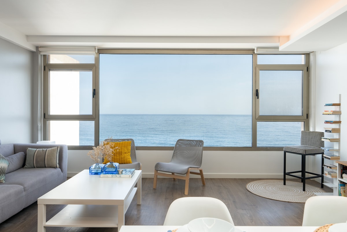 A spacious living area features a large window providing an unobstructed view of the Atlantic Ocean. A light-colored sofa is positioned near the window, with a minimalist coffee table in front. A comfortable chair and decorative elements enhance the inviting space.