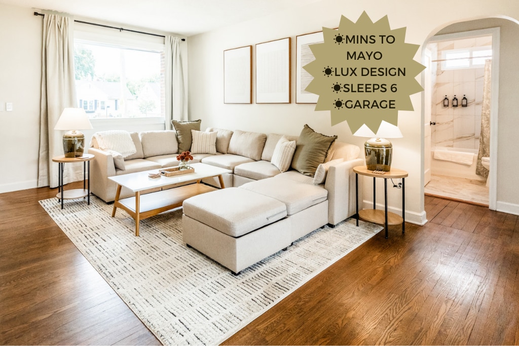 A spacious lounge area is highlighted by a light-colored sectional sofa, adorned with neutral and textured pillows. A sleek coffee table sits atop a soft area rug, enhancing the inviting atmosphere. An archway leads to a bright bathroom with modern fixtures, contributing to the home's modern design.