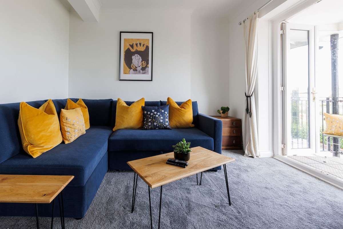 A light-filled living area is presented with an L-shaped blue sofa adorned with yellow cushions. Two wooden side tables are positioned in front of the sofa, alongside a small decorative plant. Glass doors open to a balcony, enhancing the space's airy feel.