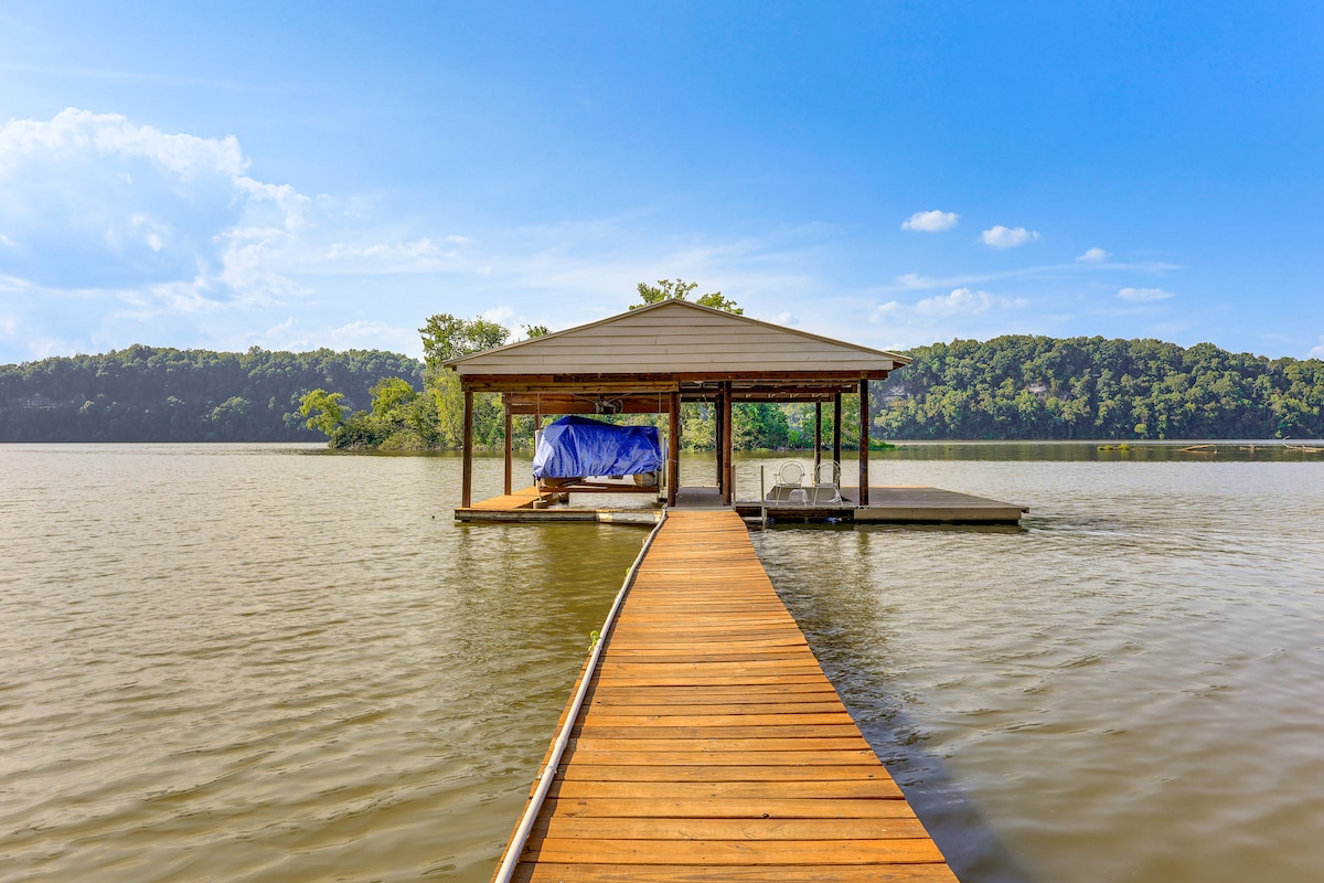 A wooden dock extends over the water, leading to a covered structure that accommodates a boat. Lush greenery borders the lake, and the calm water reflects the blue sky and clouds, enhancing the tranquil waterfront setting.