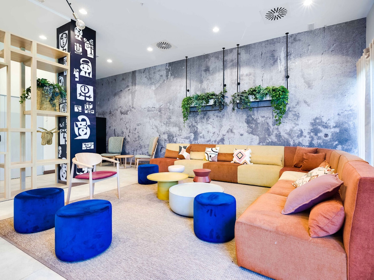 A communal lounge area features a curved sectional sofa in mixed colors and a variety of round stools in vibrant shades. Lush greenery is positioned on a modern accent wall, contributing to the inviting environment. Natural light filters through large windows, enhancing the spacious design.