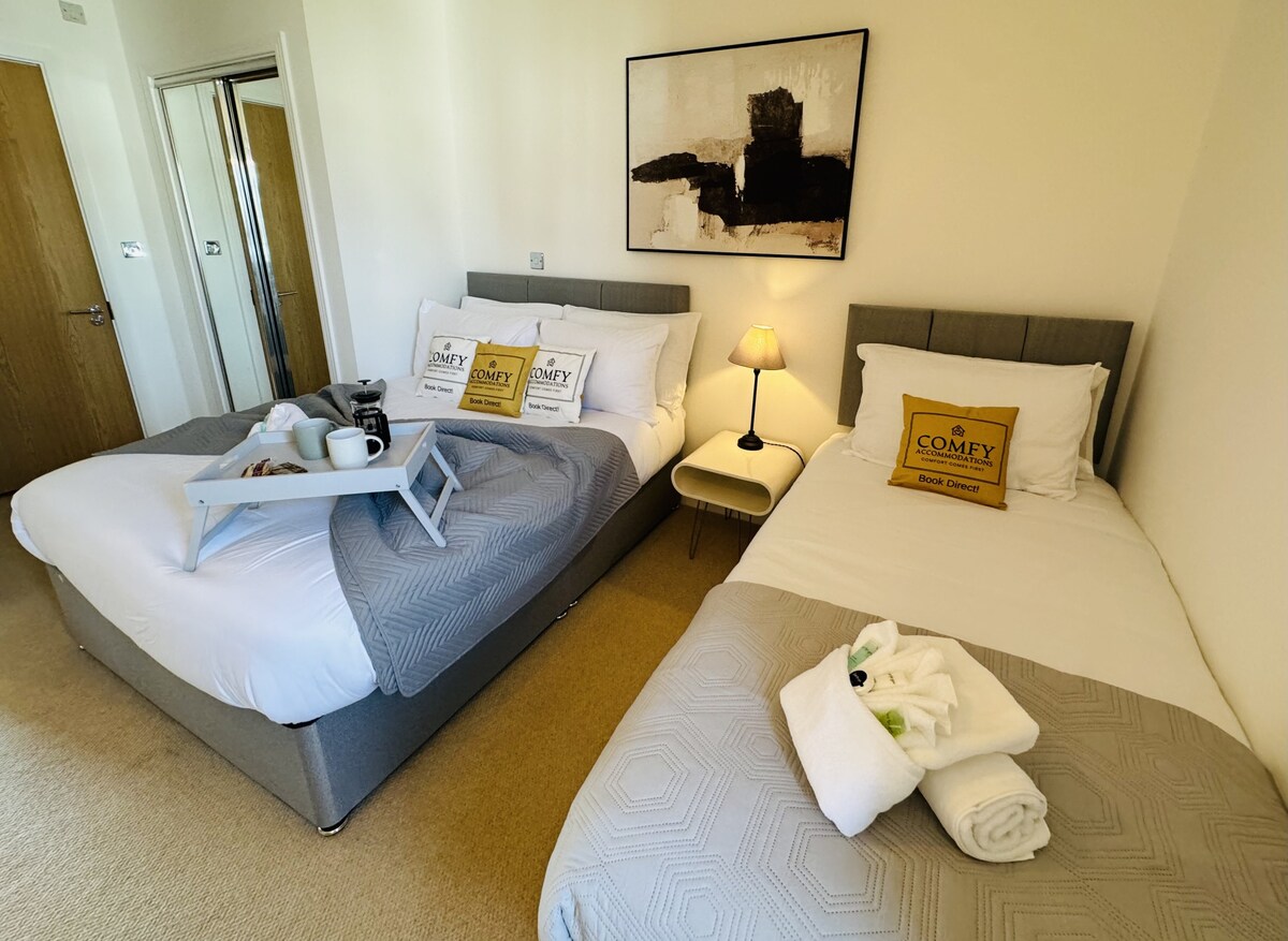 A well-arranged sleeping area features two beds, one double and one single, dressed in crisp linens. A small bedside lamp provides soft lighting, and a tray with breakfast items rests on the double bed, creating a welcoming atmosphere. Towels are neatly placed at the foot of the single bed.