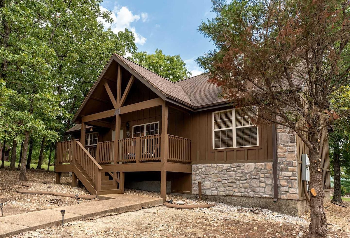 A charming cabin exterior features a combination of dark wood and stone accents. A covered porch with railings is visible, along with steps leading to the entrance. Surrounding trees provide a natural setting, enhancing the lodge's tranquil ambiance.