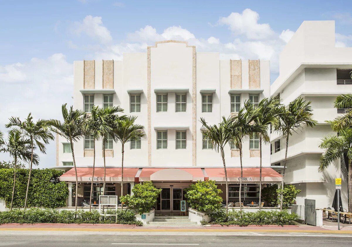 The hotel exterior showcases an Art Deco architectural style with a light color palette and distinctive geometric lines. Palm trees frame the entrance, and an awning in front adds a touch of shade. The surrounding greenery complements the building's serene atmosphere.