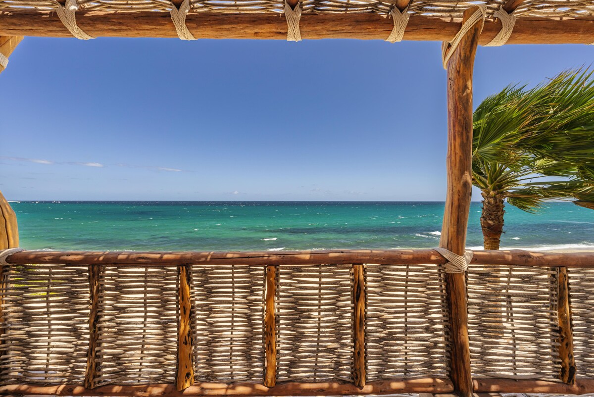 A handcrafted wooden structure frames a picturesque view of the ocean, showcasing clear blue waters meeting a clear sky. Palm trees can be seen swaying gently in the breeze, enhancing the serene beach setting.