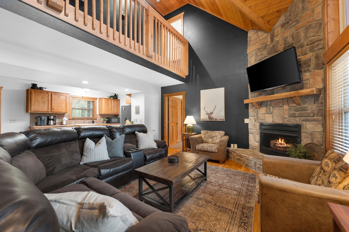 The spacious living area features a large, curved sectional sofa arranged around a coffee table. A stone fireplace is located under a wall-mounted television. Warm wood accents complement the vaulted knotty-pine ceiling, while natural light filters in through nearby windows.