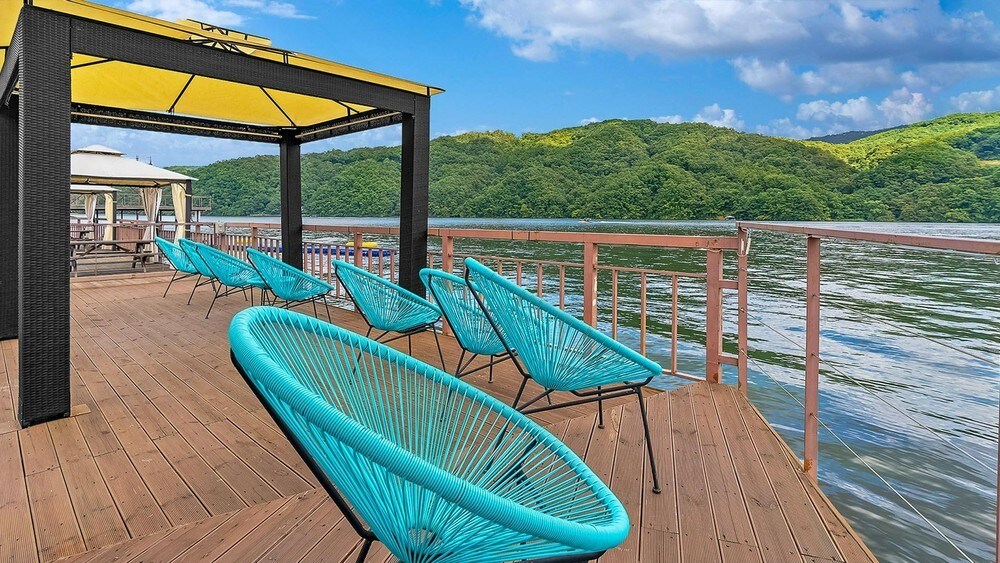 A spacious deck extends over the water, featuring multiple teal chairs arranged for relaxation. A shaded area is provided by a yellow canopy, while the backdrop showcases lush green hills under a partly cloudy sky.
