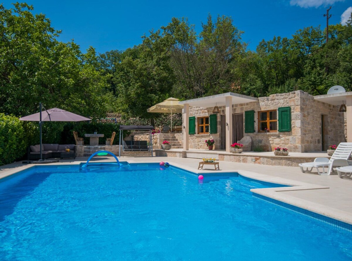 An inviting private pool is surrounded by a sun terrace, featuring lounge chairs and colorful pool floats. A stone building with green shutters and vibrant flowers is visible alongside shaded seating under an umbrella, set amidst a lush garden backdrop with trees and blue skies.