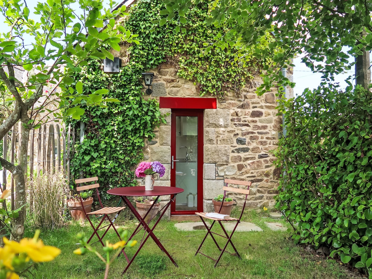 A small patio area is surrounded by green foliage and blooming flowers. A round red table with two matching chairs is positioned in front of a stone wall featuring climbing plants. A vase with flowers is centered on the table, adding a touch of color to the outdoor space.