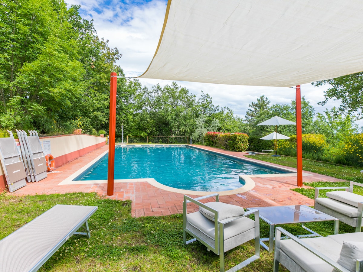 A tranquil outdoor pool area is surrounded by greenery and features comfortable lounge furniture. The pool is enclosed by a gentle slope and includes a sunshade for shade. A set of lounge chairs and a glass table are positioned nearby, enhancing the relaxing atmosphere.