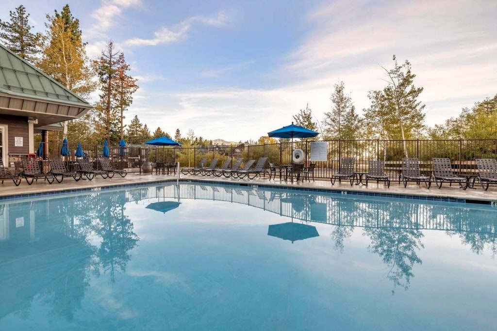 An outdoor pool is surrounded by a spacious deck, featuring lounge chairs and shaded umbrellas. Lush trees provide a natural backdrop, and clear blue waters reflect the sky. The area is designed for relaxation, with easy access to the lodge amenities nearby.
