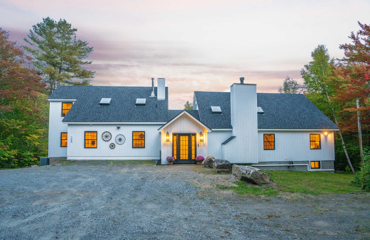 A large modern farmhouse is set against a backdrop of colorful trees at dusk. The facade features multiple windows glowing with warm light, inviting guests inside. The entrance is framed by decorative elements and a stone pathway, leading into a cozy and welcoming atmosphere.