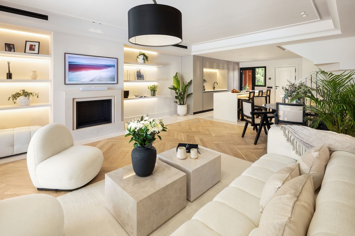 A contemporary living room features light wood flooring and a neutral color palette. Soft, rounded furniture is arranged around a low, square coffee table. A television is mounted above a sleek fireplace, while shelves adorned with plants and decor enhance the modern aesthetic.