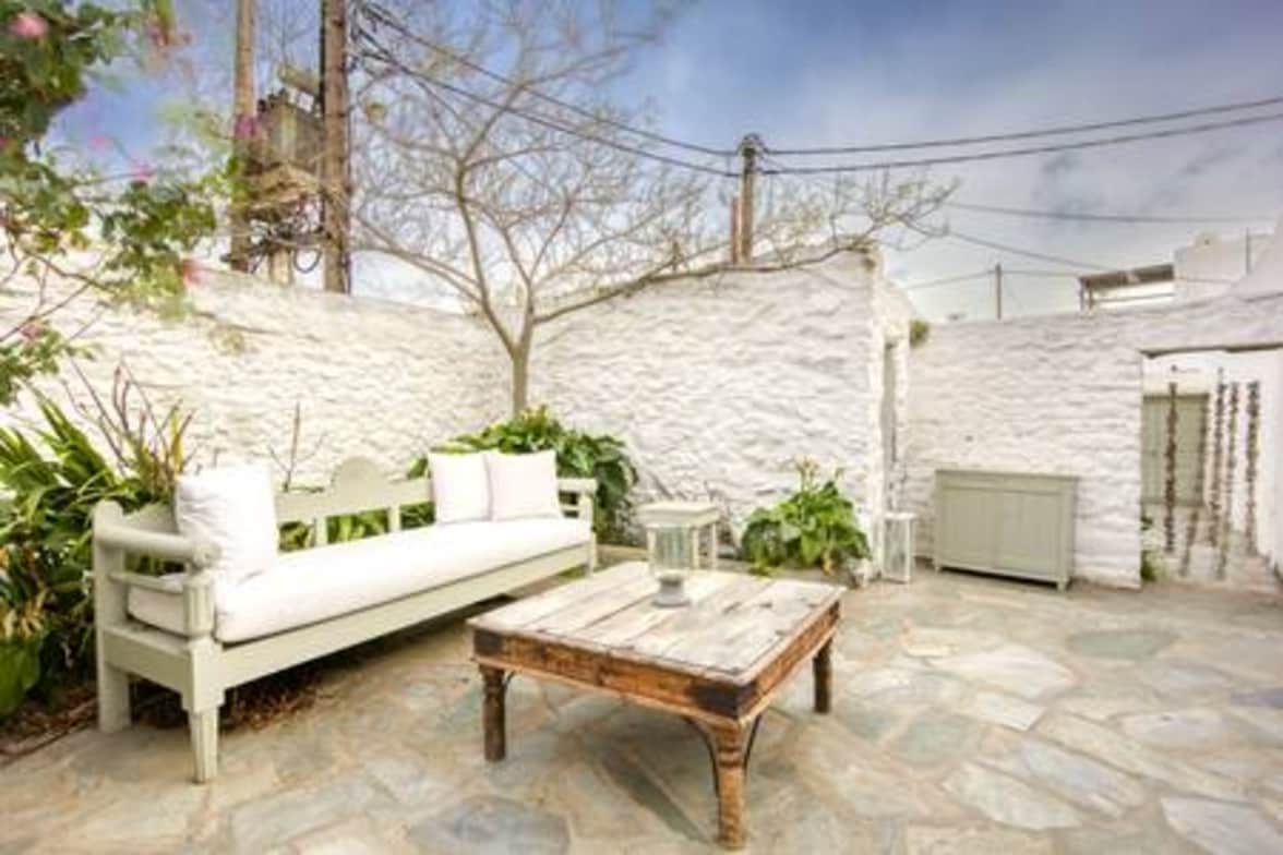 An outdoor seating area features a light-colored bench with soft cushions, accompanied by a rustic wooden table. Surrounding the patio, lush greenery adds a touch of nature against the whitewashed walls, creating a serene space for relaxation.