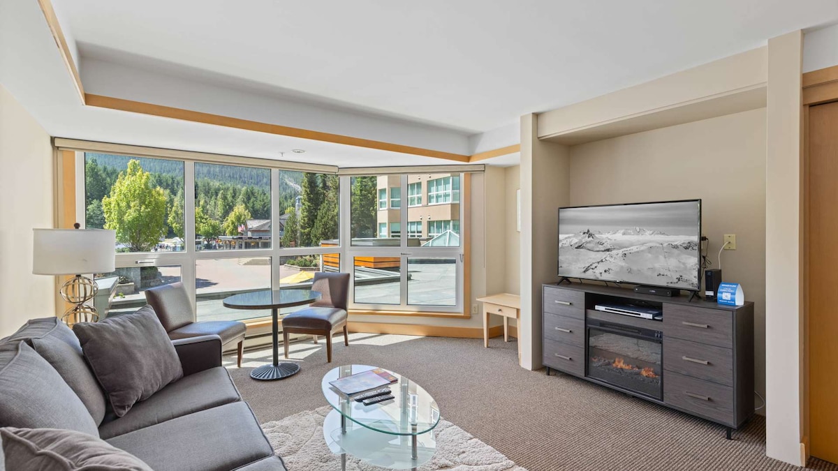 A cozy living area is presented with large windows that offer views of the surrounding landscape. A comfortable sofa and a glass coffee table create a welcoming space. A television is mounted above a contemporary fireplace, enhancing the room's inviting atmosphere.