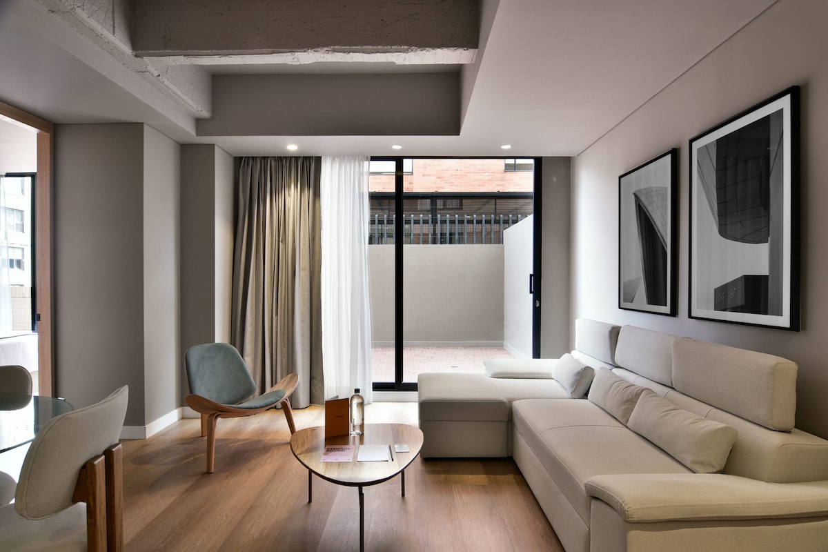 A modern living area is highlighted by a corner sofa and a glass coffee table. Floor-to-ceiling windows allow natural light to fill the room, showcasing neutral tones and minimalist artwork. A cozy chair is positioned nearby, providing an inviting spot for relaxation.