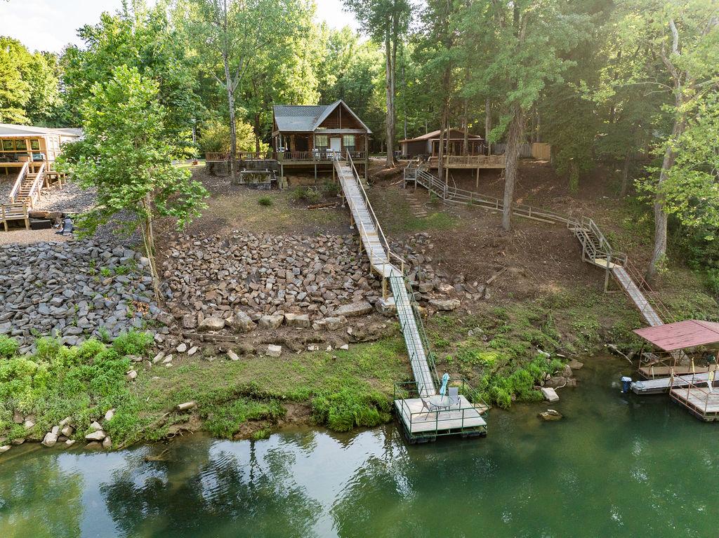 The cabin is positioned along the Little Red River, featuring a private dock accessible by a long boardwalk. A rocky shoreline and lush greenery surround the area, with another cabin visible nearby, creating a tranquil riverfront setting.