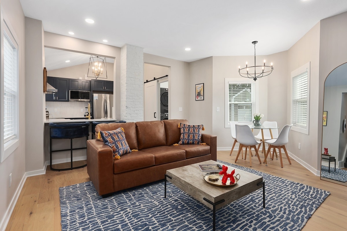 A modern living space is shown, featuring a brown leather sofa adorned with patterned cushions. A wooden coffee table sits on a textured blue rug, while a dining area with white chairs and a round chandelier is visible in the background. Natural light enters through multiple windows.