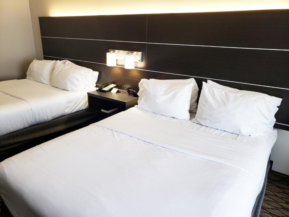 A spacious room showcases two neatly made beds with white linens and a dark wooden headboard. Soft lighting from adjacent wall fixtures creates a calm ambiance, while a bedside table holds a phone. A dark bench is positioned at the foot of the beds.
