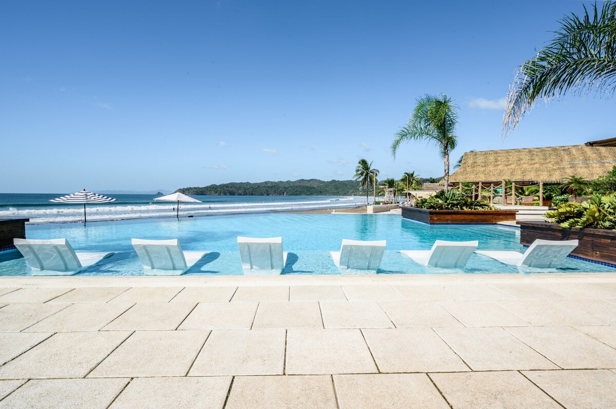 An outdoor infinity pool is highlighted, featuring several loungers positioned along the water's edge. Lush palm trees surround the area, offering a tropical ambiance. The beach and ocean extend beyond the pool, with gentle waves visible in the distance under a clear blue sky.