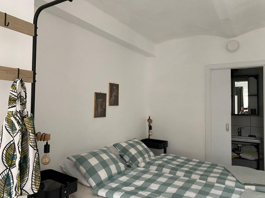 A comfortable double bed is adorned with checkered bedding, positioned against a neutral wall. Two framed pictures are hung nearby, and a small table with a lamp stands beside the bed. A doorway visible in the background leads to another area.