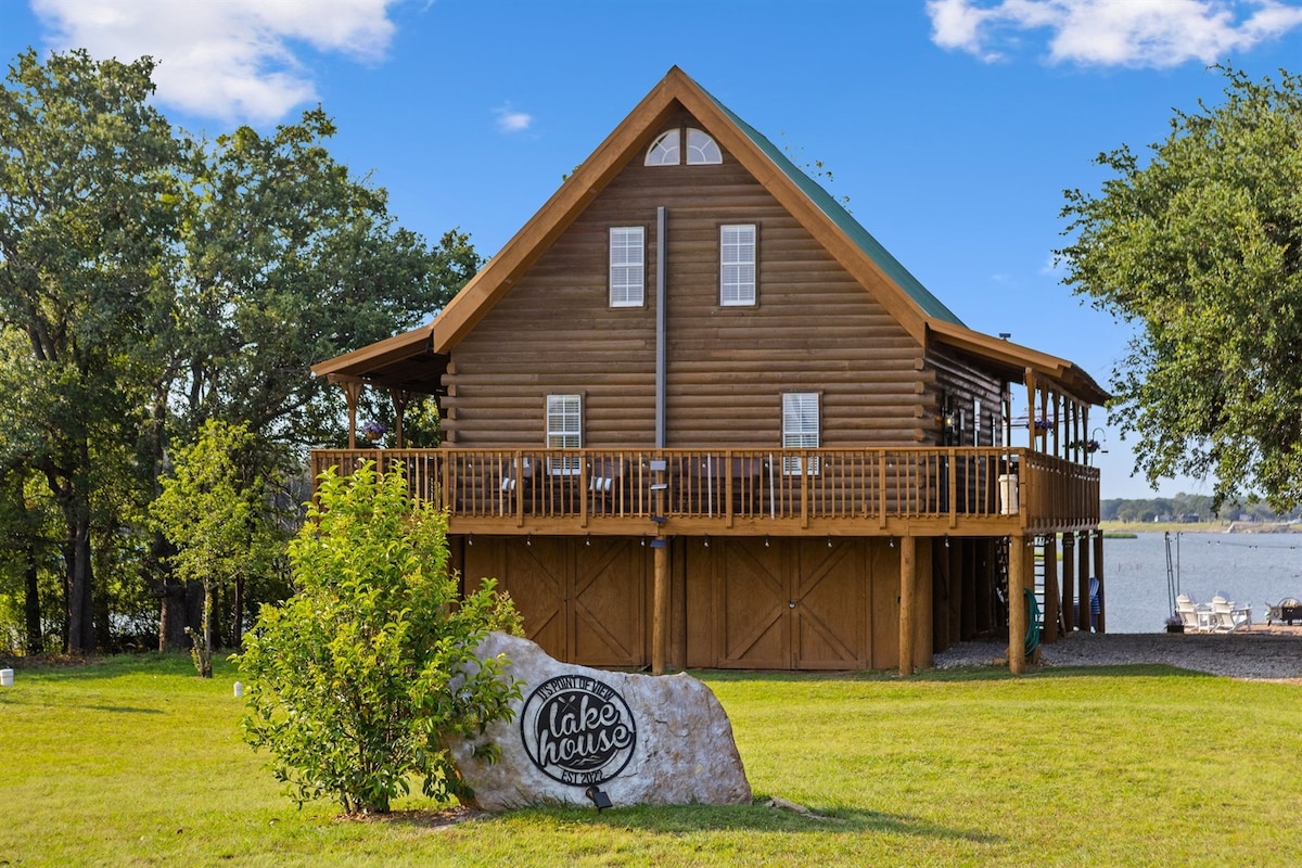 A lifted log cabin home with a wraparound deck is set against a backdrop of lush greenery. The exterior is structured with natural wood and a green roof, accented by a large stone displaying the word 'lake house.' The serene lake can be seen in the distance.