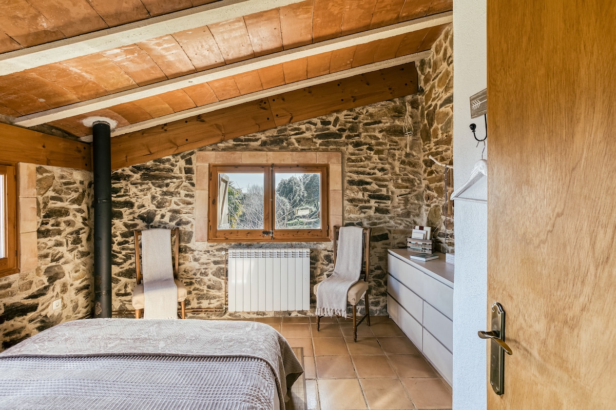 A cozy bedroom features exposed stone walls and wooden beams, enhancing the rustic charm. A spacious bed is complemented by two armchairs positioned near a window that offers views of the outdoors. A dresser provides additional storage, creating a comfortable and serene atmosphere.