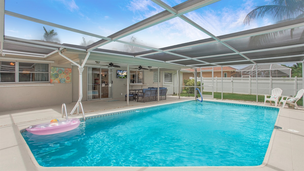 A private pool area is featured, surrounded by a screened enclosure for comfort. Seating is provided near the water, with a cozy lounge set and a flat-screen TV visible in the background. A colorful pool float rests in the inviting blue water under a clear sky.