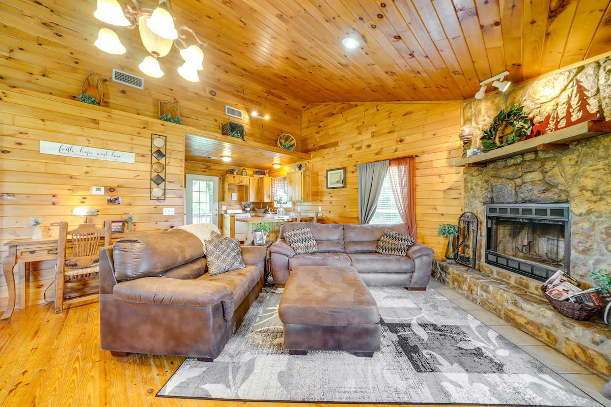 A spacious living area features wooden walls and ceiling, complemented by a stone fireplace. Comfortable brown sofas, adorned with patterned cushions, invite relaxation. A light fixture hangs above, and natural light filters through large windows, illuminating the adjacent dining area and kitchen.