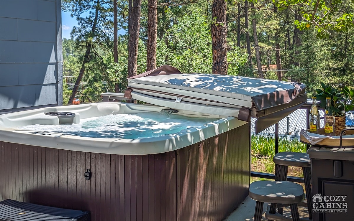 A private hot tub is shown with its cover partially lifted, revealing bubbling water. Surrounding the tub are wooden stools, and the lush greenery of trees creates a serene atmosphere.