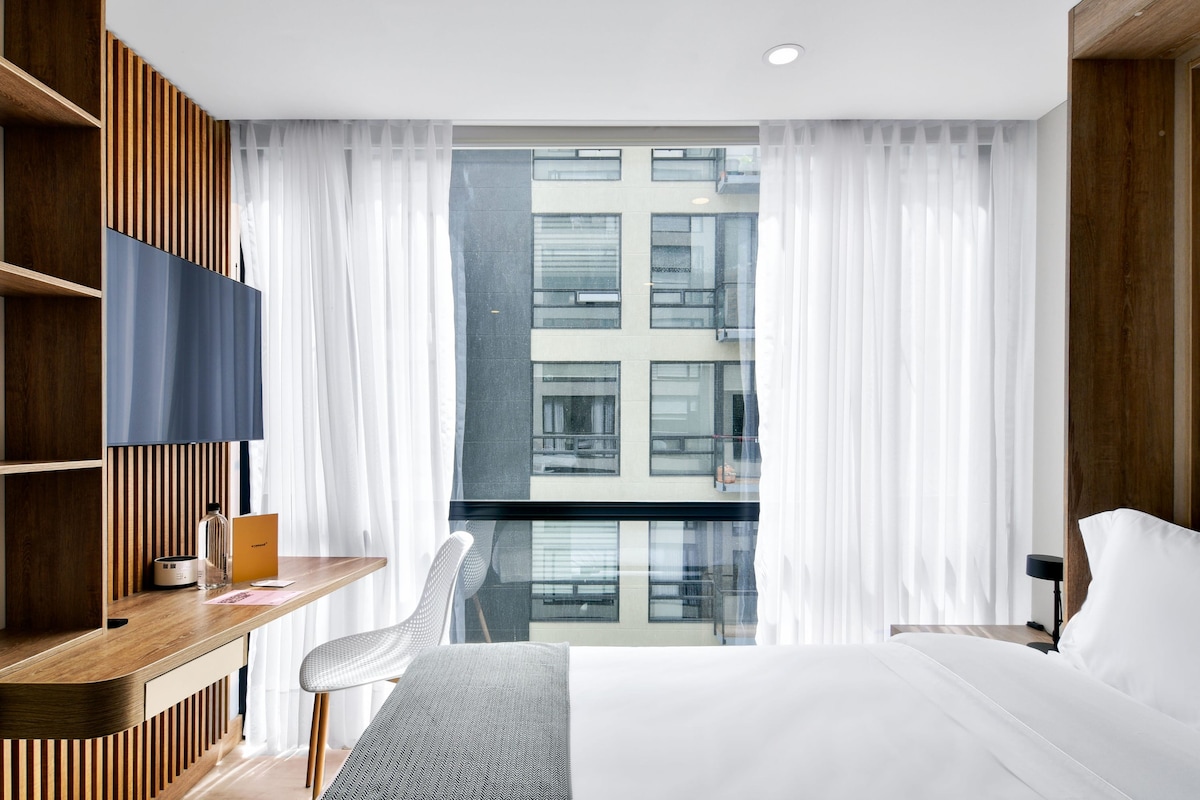 A modern room features a spacious bed with crisp white linens, complemented by a wooden accent wall and a desk area. Large windows dressed in sheer white curtains provide natural light and a view of the building outside.