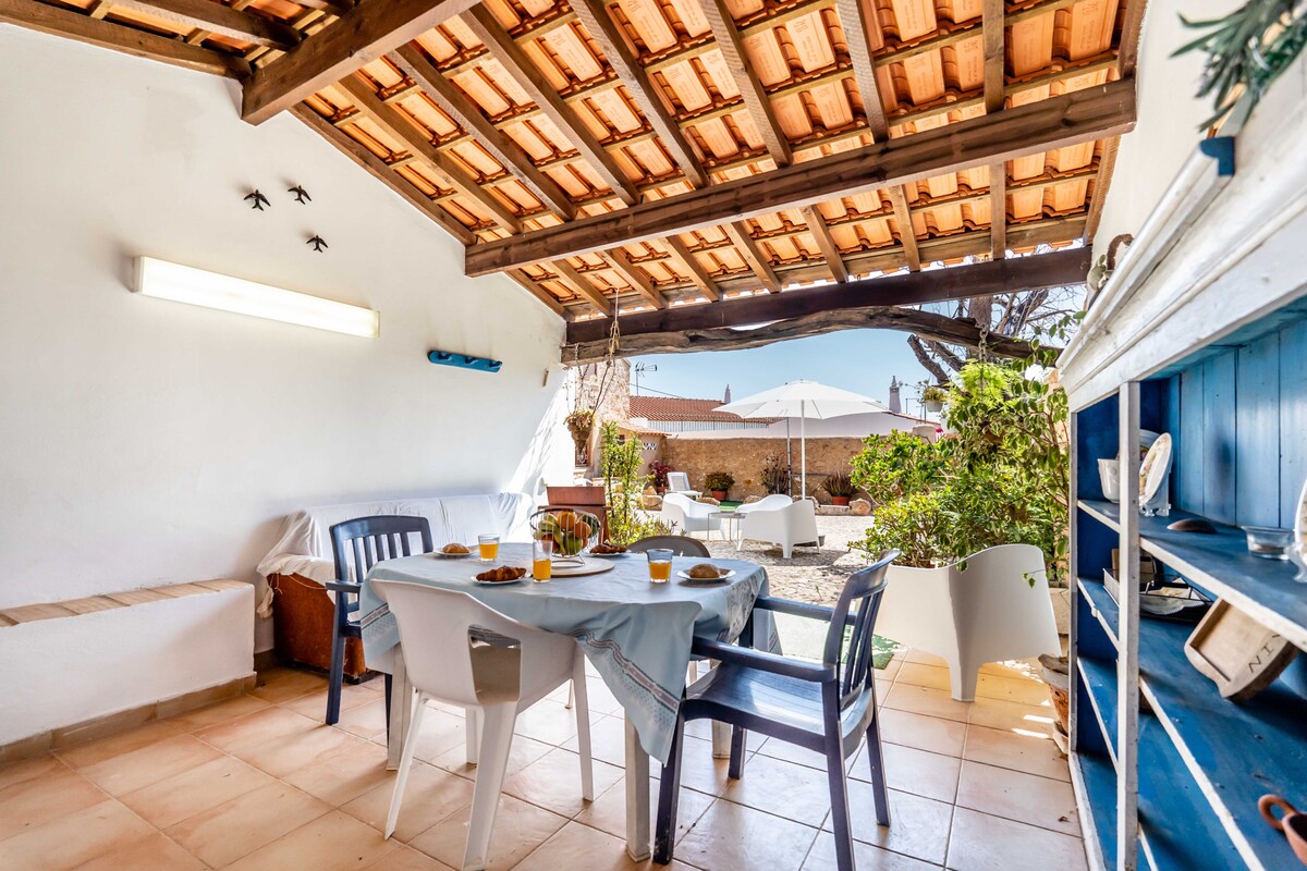 An outdoor dining area features a table set for meals, surrounded by chairs. A shaded structure with wooden beams provides cover, while potted plants and a glimpse of garden seating are visible in the background. Soft lighting is installed along the wall.