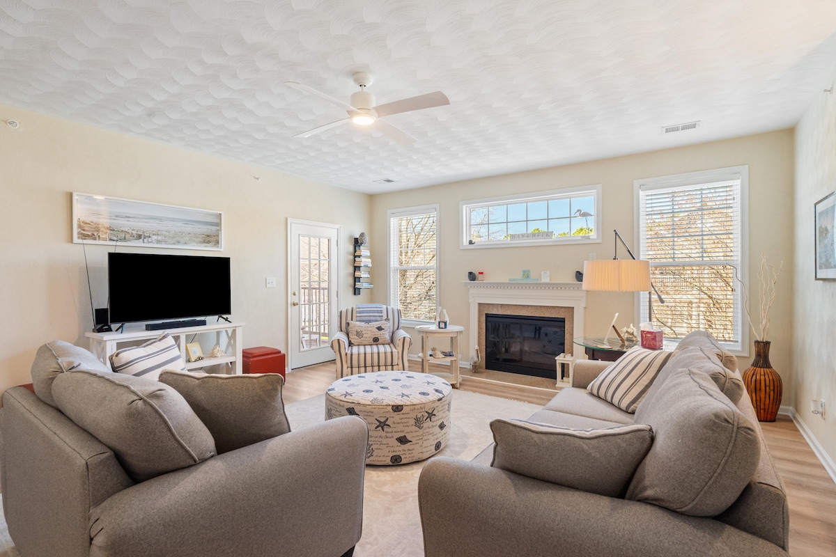 A bright living room features a fireplace at the center, framed by two large windows allowing natural light. Comfortable gray sofas are positioned around a round, patterned ottoman, and a flat-screen TV is mounted on a light-colored wall.