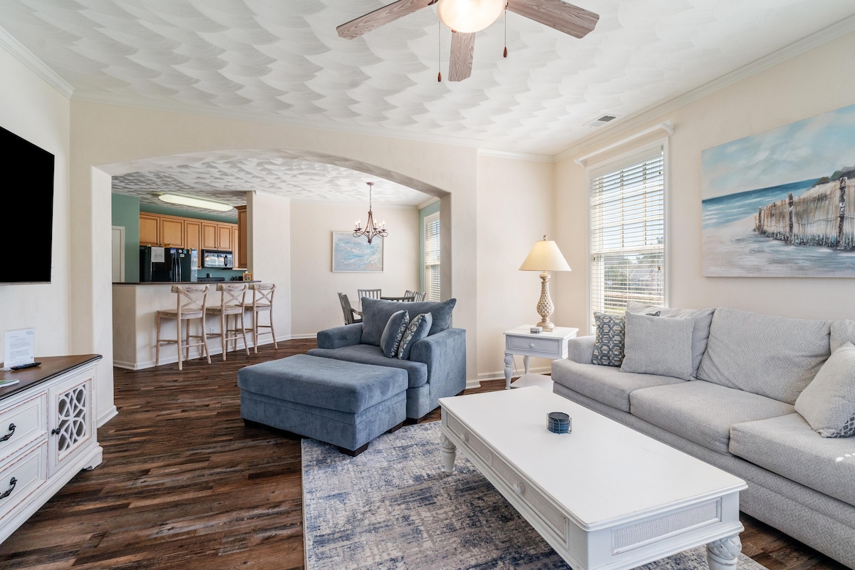 The living area features a spacious sectional sofa complemented by a matching chaise lounge. A large flat-screen TV is mounted on the wall, while a decorative coffee table occupies the center. Natural light fills the room through multiple windows, enhancing the coastal decor.