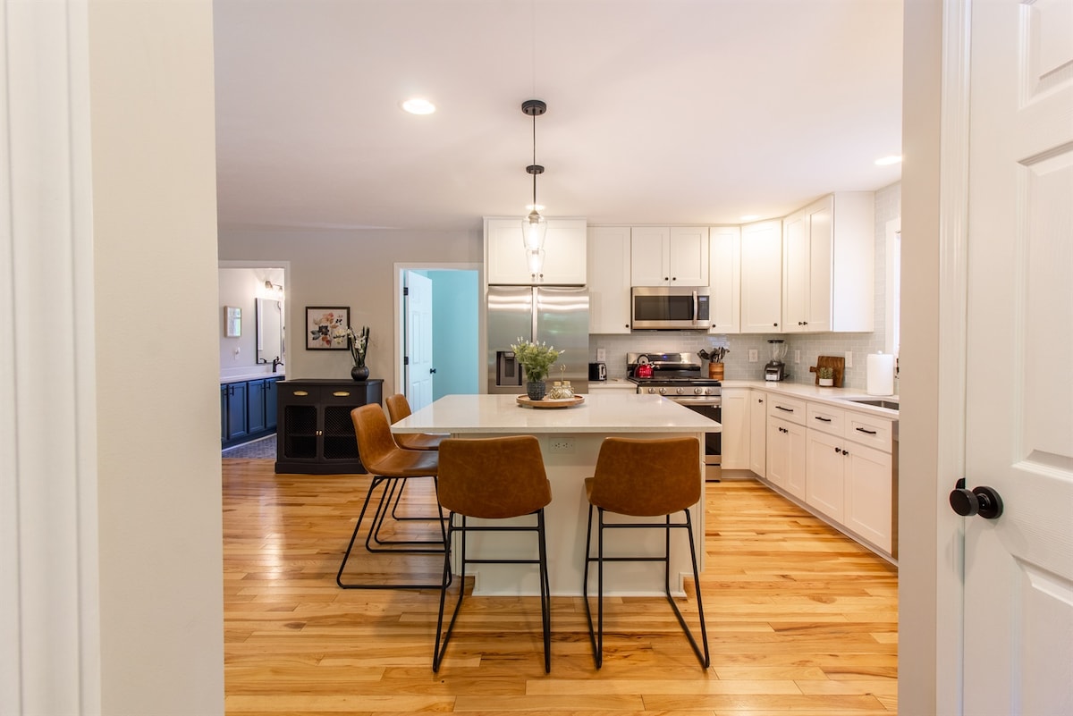 The open kitchen showcases modern cabinetry and stainless steel appliances. A central island with seating for three is highlighted by warm wood flooring. Natural light brightens the space, and a glimpse of the living area is visible through the doorway.