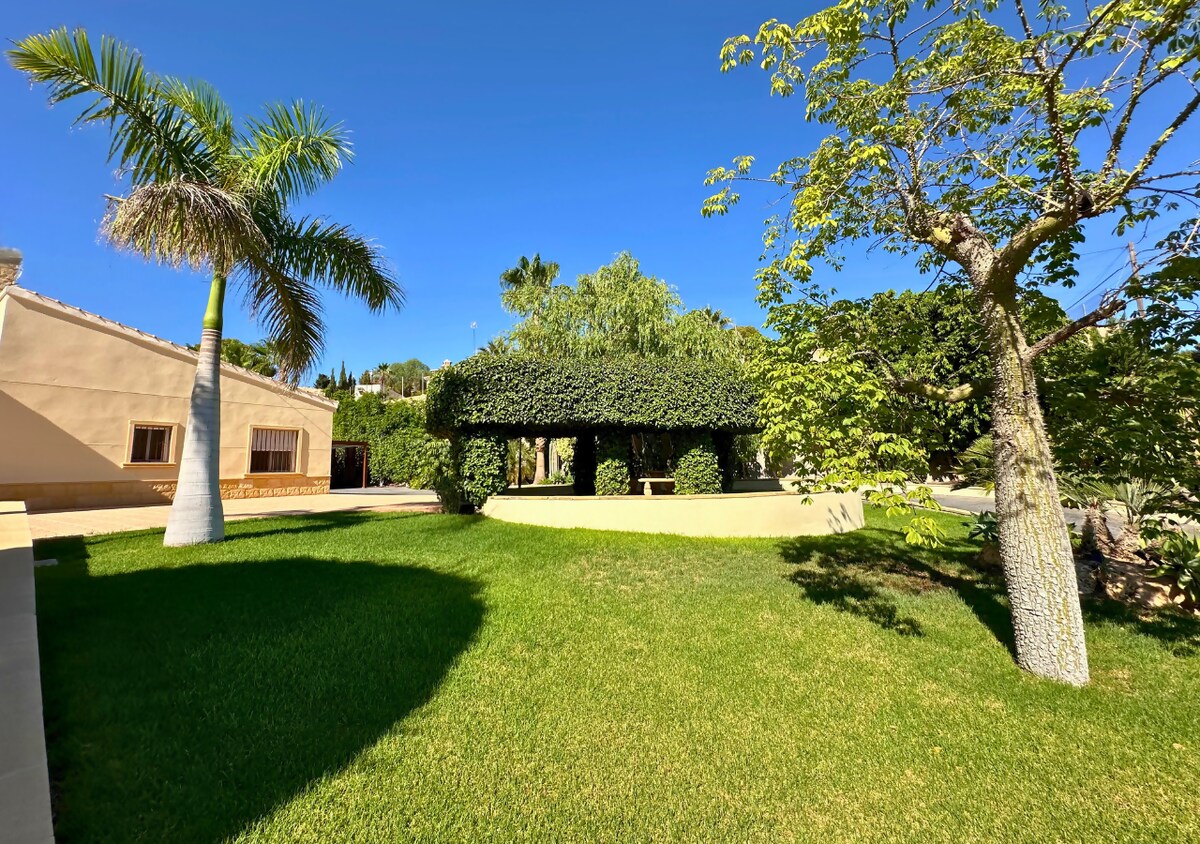 Lush green grass surrounds a shaded outdoor seating area covered in greenery, providing a cozy space for relaxation. Palms and other trees are visible, enhancing the serene garden atmosphere of the villa. A clear blue sky adds brightness to the tranquil scene.