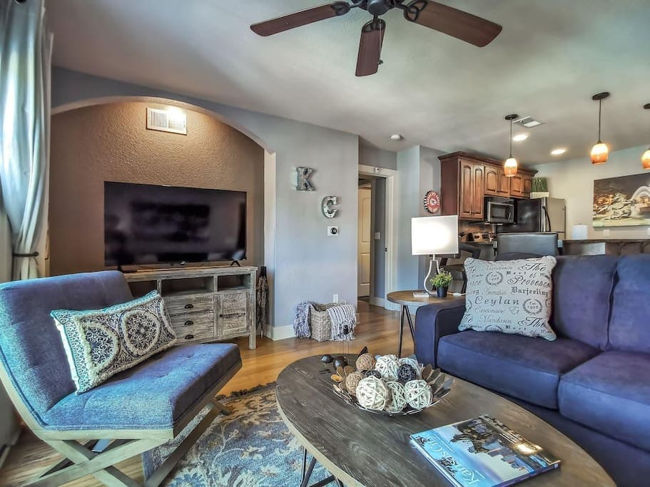 The living area is furnished with a cozy sofa and an accent chair, complemented by a round coffee table. A large television is mounted on the wall, while a warm wooden floor adds character. Soft lighting from multiple sources creates an inviting ambiance.